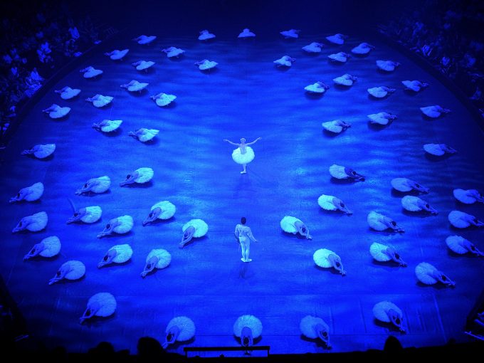 A dancer in the role of Odette from Swan Lake and a dancer in the role of Prince Siegfried are facing each other at the centre of a round stage filled with blue light. Odette is wearing a white tutu and headdress while Siegfried is wearing a white top and white tights. Around them, a large group of dancers playing the white swans are shown laying on the ground in perfect unison, all wearing white tutus.