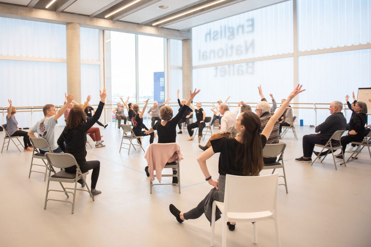 A group of people living with Parkinson's, and their carers, are taking a dance class in a bright studio. They are sat in a big circle, on chairs.