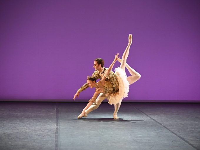 A dancer in the role of Prince Désiré from The Sleeping Beauty is holding a dancer in the role of Aurora who is leaning forward with her legs up in the air in a beautiful ballet pose. The Prince is wearing a golden top and white tights while Aurora is wearing a golden and white tutu. They are on a stage with a violet background.