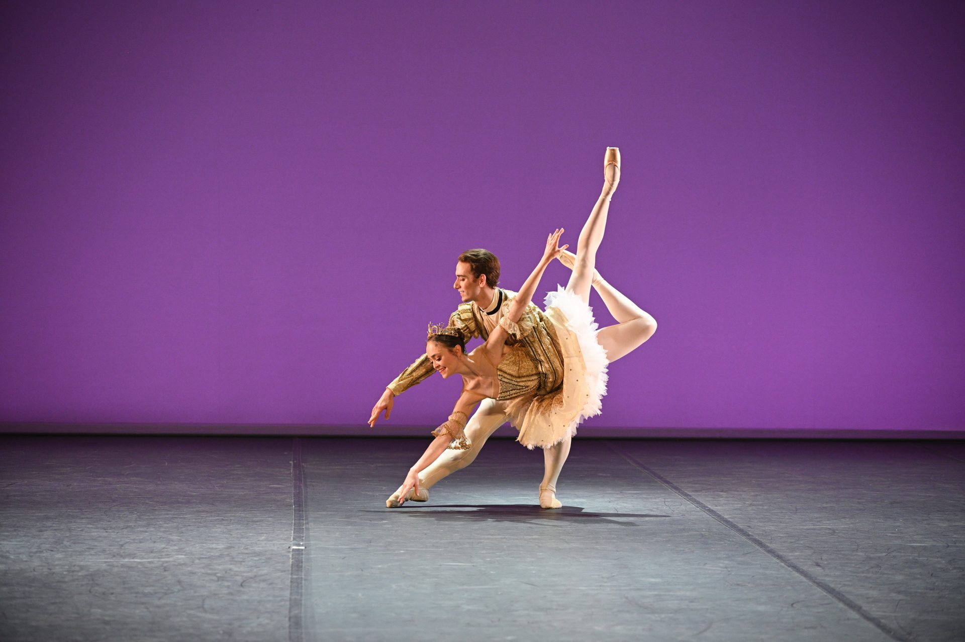 A dancer in the role of Prince Désiré from The Sleeping Beauty is holding a dancer in the role of Aurora who is leaning forward with her legs up in the air in a beautiful ballet pose. The Prince is wearing a golden top and white tights while Aurora is wearing a golden and white tutu. They are on a stage with a violet background.