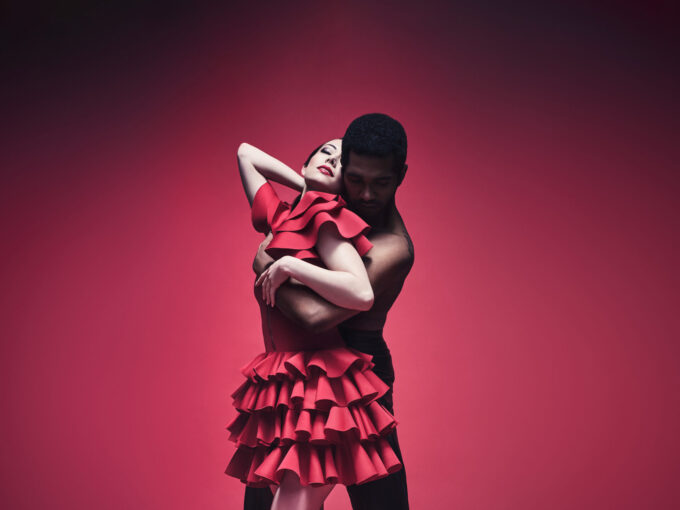A dancer playing the role of Carmen and a dancer playing the role of Don Jose are shown in a sensual embrace over a dark red background. Carmen is wearing a short red dress with frills while Don Jose is wearing dark trousers.