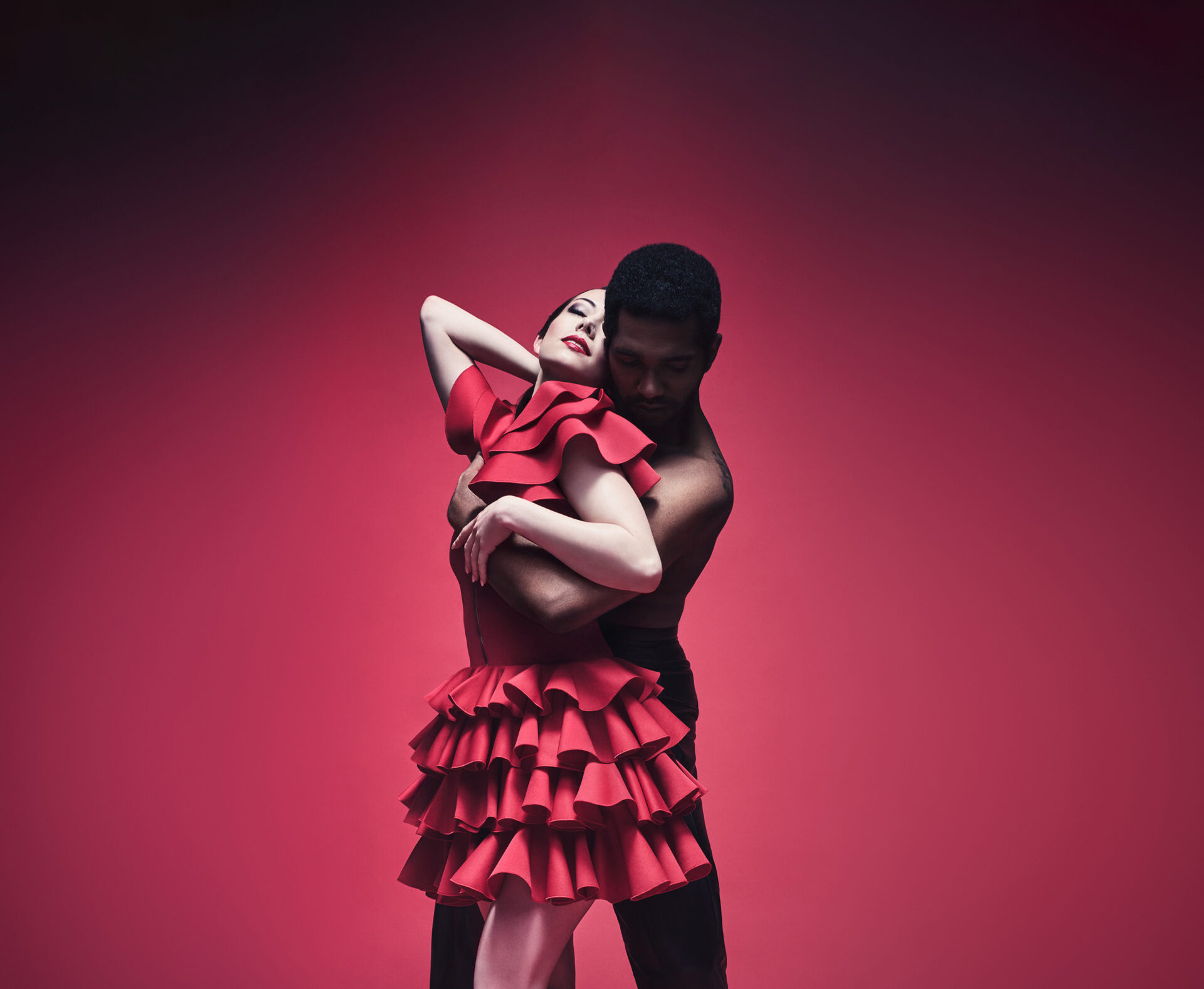 A dancer playing the role of Carmen and a dancer playing the role of Don Jose are shown in a sensual embrace over a dark red background. Carmen is wearing a short red dress with frills while Don Jose is wearing dark trousers.