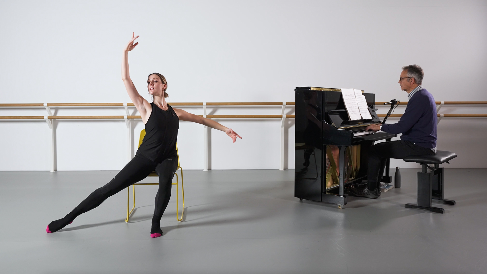 A dance tutor does a port de bras while sitting on a chair. A pianist is in the background.