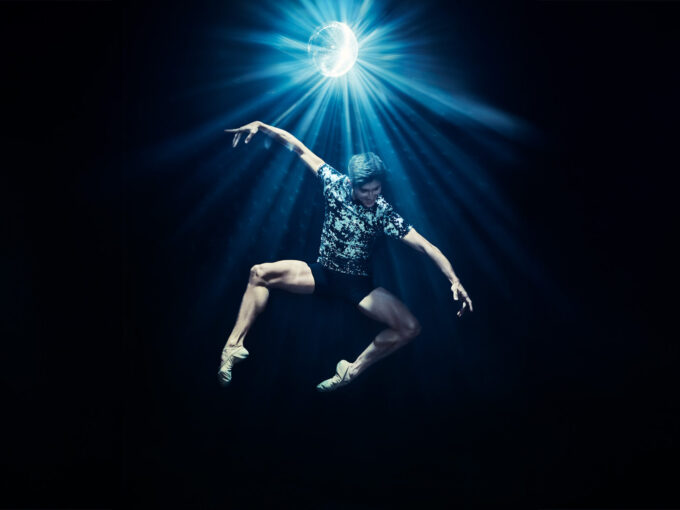 A dancer is jumping in a pas de chat, seemingly floating over a dark background lit only by an exciting disco ball. He's smiling and wearing a textured black and white shirt and black shorts.