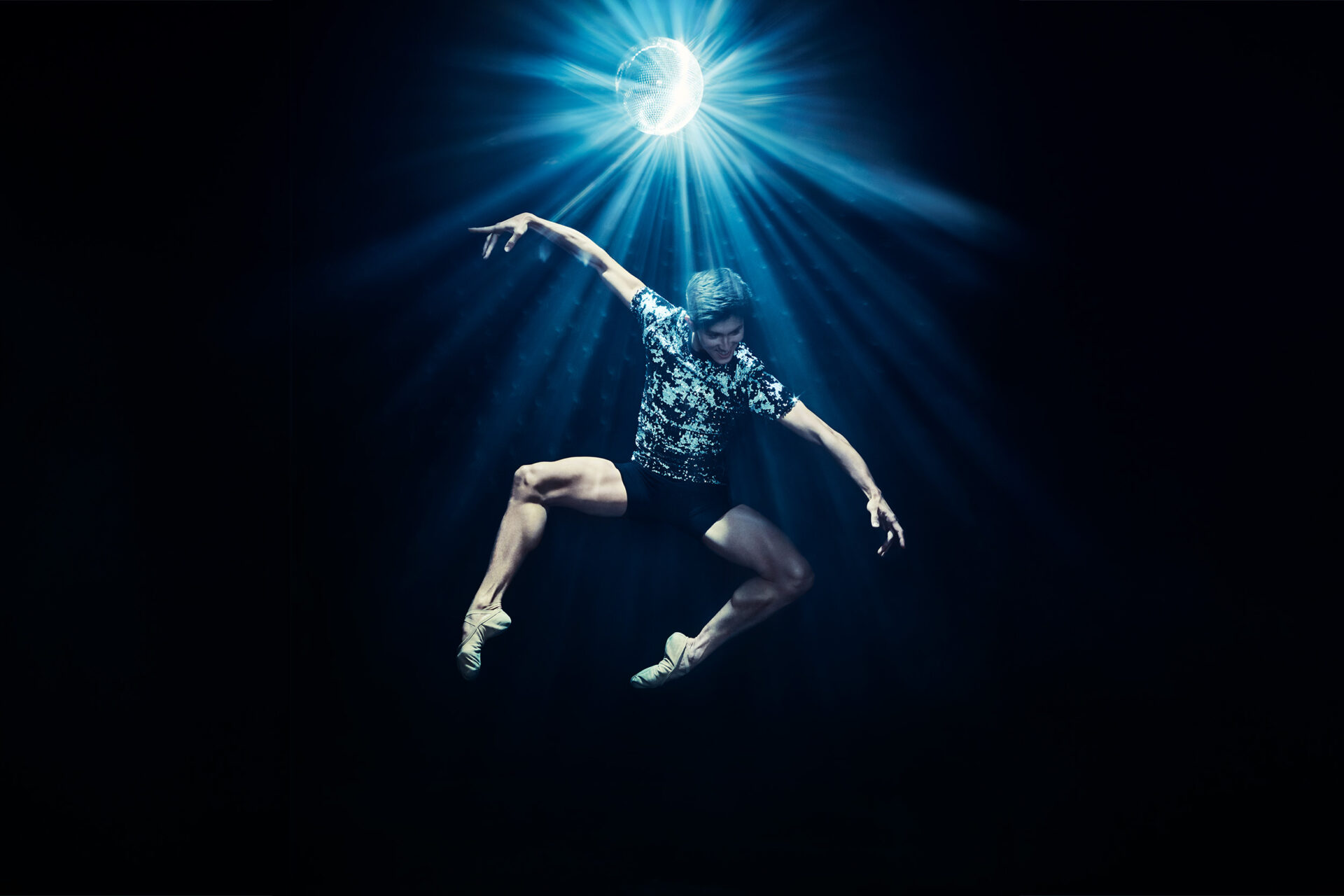 A dancer is jumping in a pas de chat, seemingly floating over a dark background lit only by an exciting disco ball. He's smiling and wearing a textured black and white shirt and black shorts.
