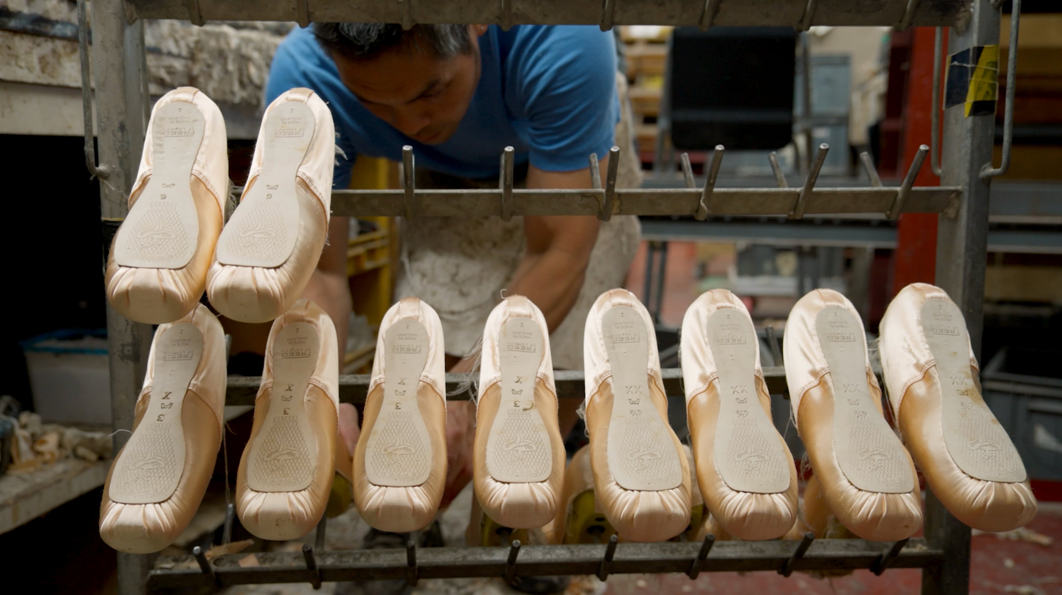 A person in a blue shirt working at a workbench in a shoe manufacturing facility, inspecting a row of ten pale pink pointe ballet shoes arranged on metal pegs,