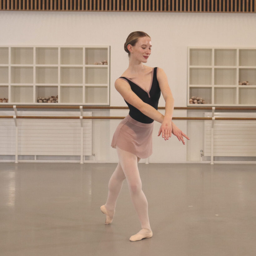 A dancer stands in the centre in a tendu in plie while her arms are laid over each other for a Nutcracker class