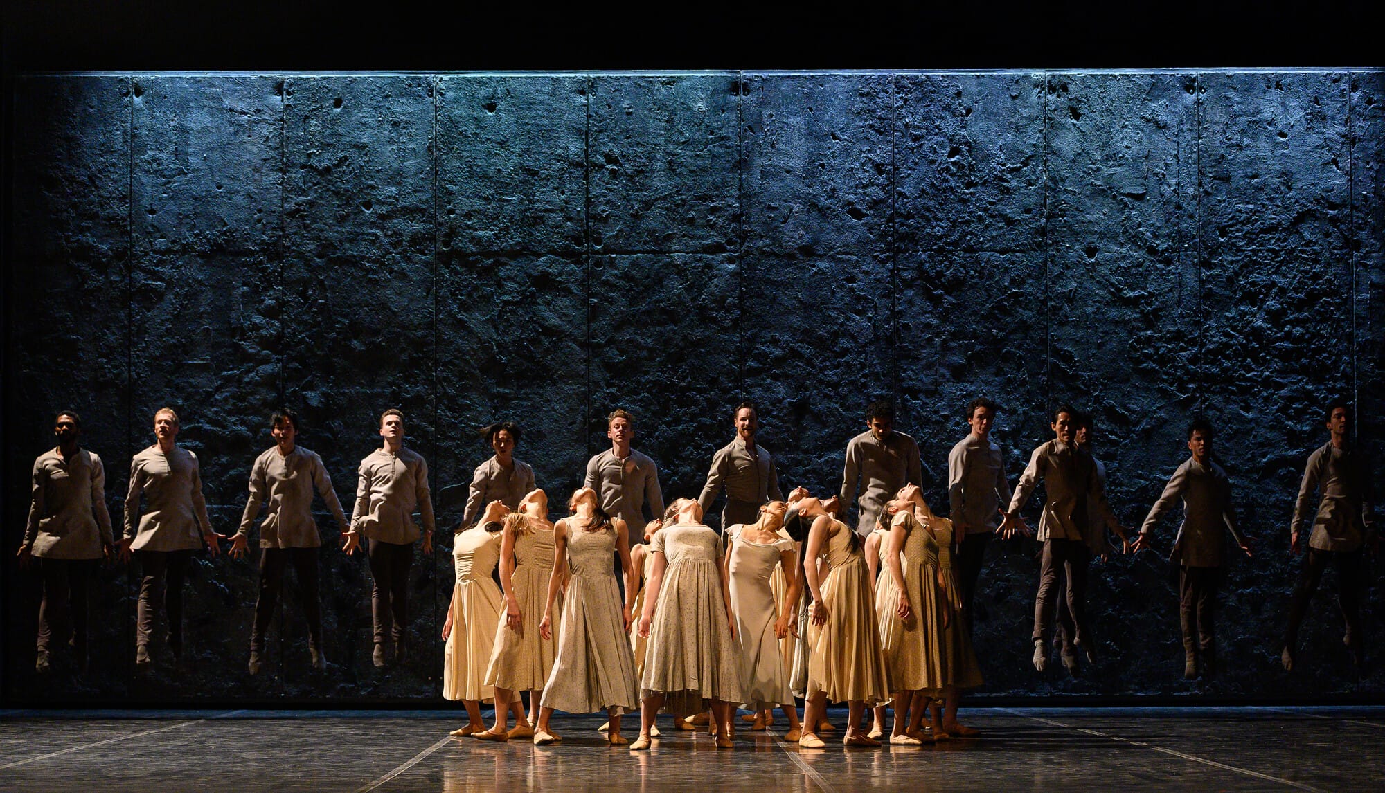 A group of ballet dancers from Akram Khan’s Giselle is divided into two distinct formations on stage. In the foreground, female dancers in cream-colored dresses gather tightly together, their heads thrown back in unison. Behind them, a line of male dancers dressed in grey coats hovers midair against a dark, textured backdrop.