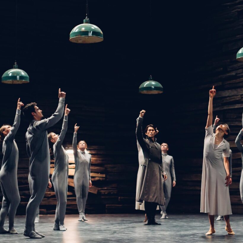 A group of dancers is shown pointing upwards towards the ceiling, four lamps hang above them