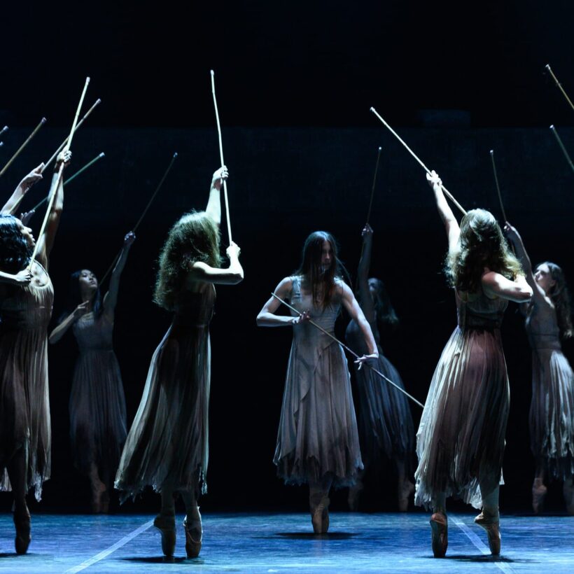 Ballet dancers, dressed in flowing, tattered beige costumes, perform on stage in an intense, ritualistic scene from Akram Khan’s Giselle. They hold long wooden sticks aloft, reaching towards the sky. The dimly lit stage creates a haunting and atmospheric effect. The central figure, with long hair obscuring her face, stands out, moving with commanding strength.
