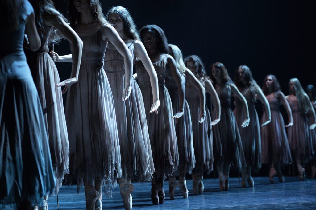 English National Ballet’s dancers performs as Wilis from Akram Khan's Giselle. The dancers stand in a line, dressed in tattered dresses, and pose on pointe with their arms extended in a menacing way. The stage is dimly lit, creating a haunting and dramatic atmosphere.