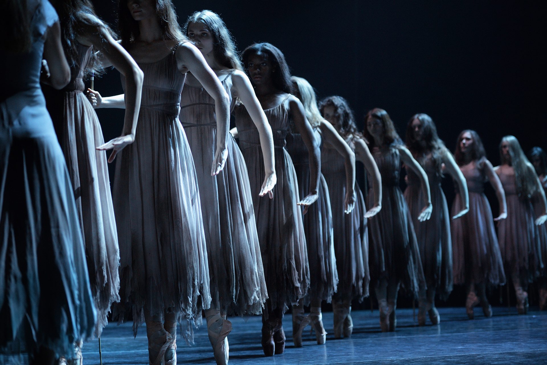 English National Ballet’s dancers performs as Wilis from Akram Khan's Giselle. The dancers stand in a line, dressed in tattered dresses, and pose on pointe with their arms extended in a menacing way. The stage is dimly lit, creating a haunting and dramatic atmosphere.