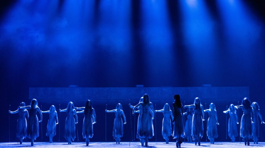 Dancers stand on pointe holding sticks, under blue spotlights, in a scene from Akram Khan's Giselle