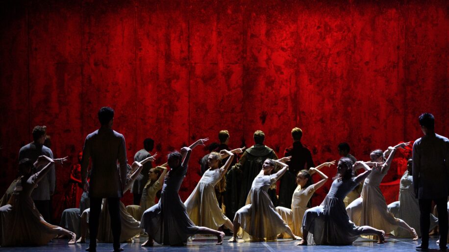 Dancers in grey dresses move in front of a large wall, lit in red, in the ballet Akram Khan's Giselle