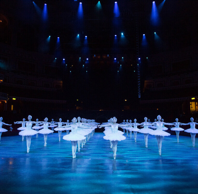 The white swans corps de ballet dancers are show across multiple lines. They are on pointe and wearing white tutus surrounded by atmospheric blue lighting