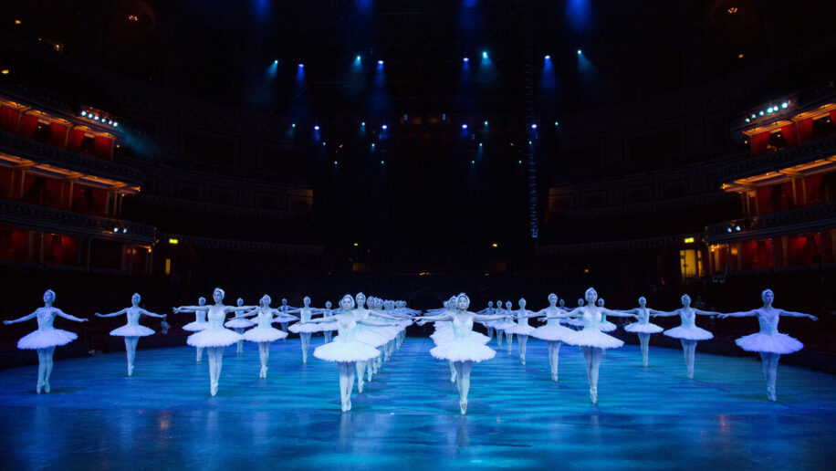 The white swans corps de ballet dancers are show across multiple lines. They are on pointe and wearing white tutus surrounded by atmospheric blue lighting