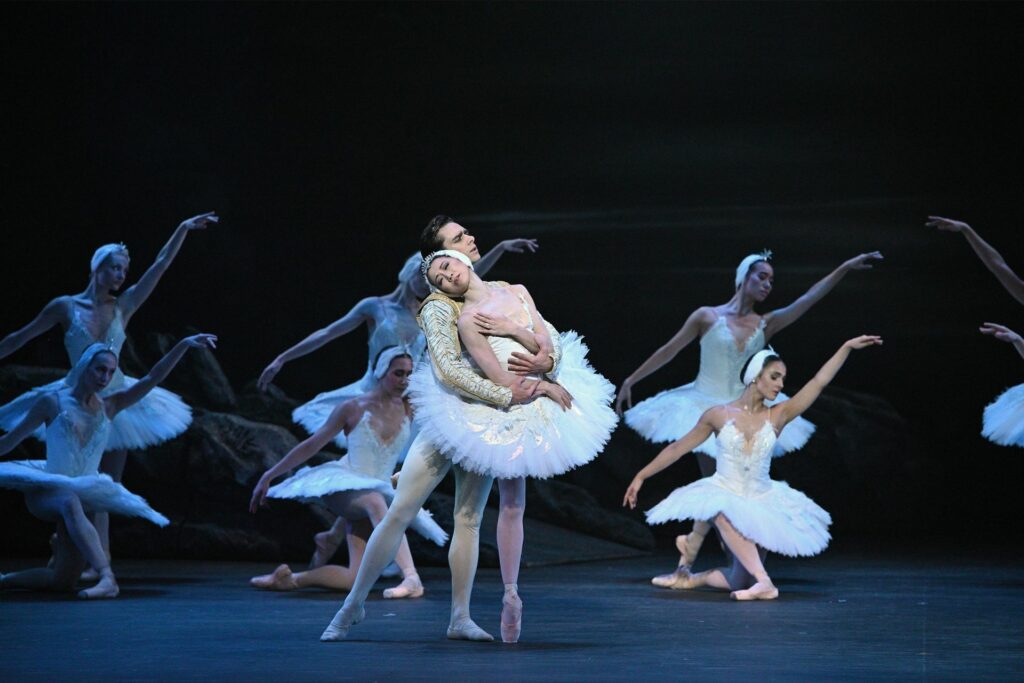 Ballet dancers performing a scene from "Swan Lake”, one of the most emotional ballets. The central dancers, portraying Odette and Prince Siegfried in elegant white costumes, are in a tender embrace, with the female dancer leaning back gracefully. Surrounding them are other ballerinas in White Swan tutus and striking delicate poses.