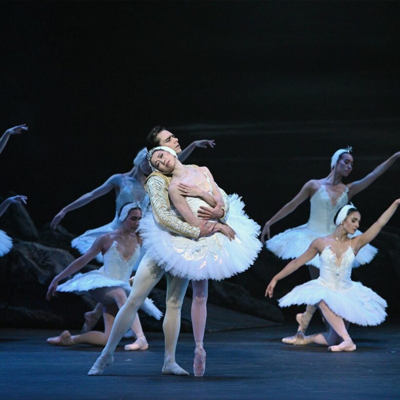 Ballet dancers performing a scene from "Swan Lake”, one of the most emotional ballets. The central dancers, portraying Odette and Prince Siegfried in elegant white costumes, are in a tender embrace, with the female dancer leaning back gracefully. Surrounding them are other ballerinas in White Swan tutus and striking delicate poses.