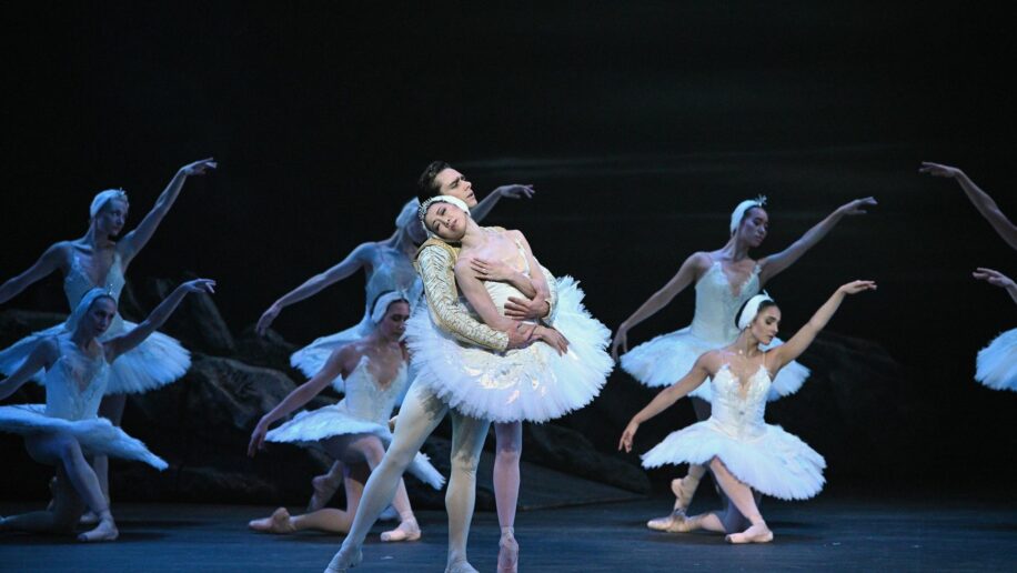 Ballet dancers performing a scene from "Swan Lake”, one of the most emotional ballets. The central dancers, portraying Odette and Prince Siegfried in elegant white costumes, are in a tender embrace, with the female dancer leaning back gracefully. Surrounding them are other ballerinas in White Swan tutus and striking delicate poses.