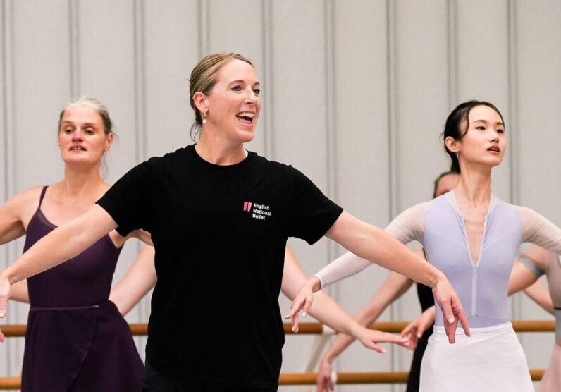 A teacher wears all black and stands at the front of a group with her arms outstretched to the side and one leg slightly behind. There are several other dancers who stand behind her, copying her movement.