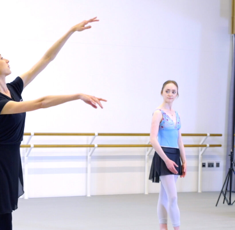 In a bright dance studio, a ballet instructor dressed in a black top, black skirt, and black leggings demonstrates a ballet pose with her arms gracefully extended and her gaze upward. In the background, a young ballerina in a light blue leotard, black skirt, and white tights stands watching attentively. On the right side of the image, a pianist sits at a piano.