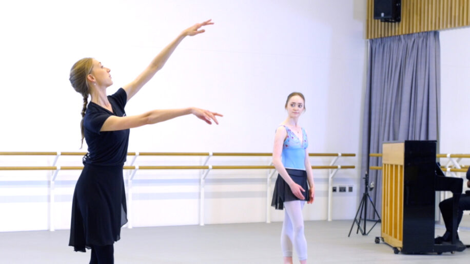 In a bright dance studio, a ballet instructor dressed in a black top, black skirt, and black leggings demonstrates a ballet pose with her arms gracefully extended and her gaze upward. In the background, a young ballerina in a light blue leotard, black skirt, and white tights stands watching attentively. On the right side of the image, a pianist sits at a piano.