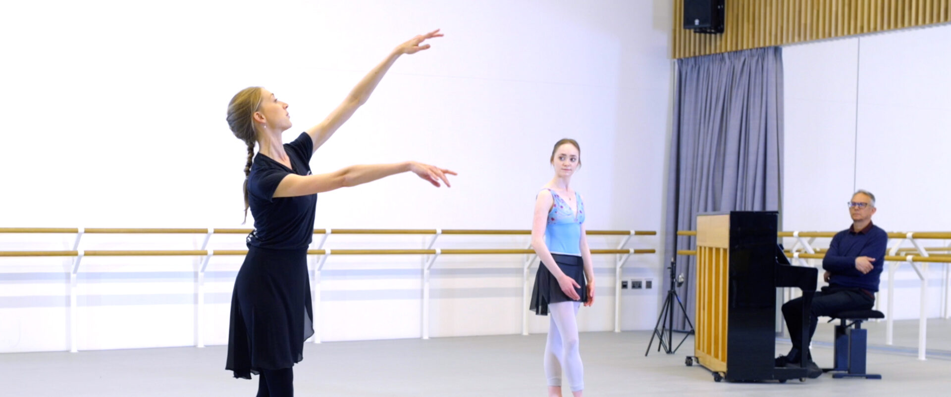 In a bright dance studio, a ballet instructor dressed in a black top, black skirt, and black leggings demonstrates a ballet pose with her arms gracefully extended and her gaze upward. In the background, a young ballerina in a light blue leotard, black skirt, and white tights stands watching attentively. On the right side of the image, a pianist sits at a piano.