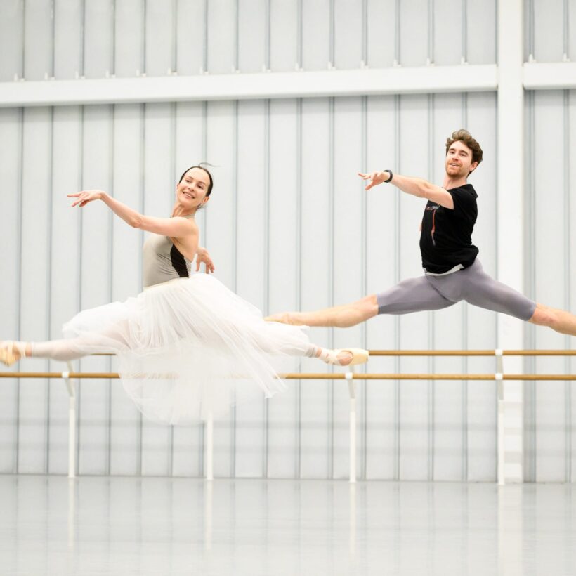 Two ballet dancers are captured mid-air in a rehearsal studio. The female dancer on the left is wearing a long, flowing white tutu and beige leotard, with her legs extended gracefully in a split leap. She has a serene expression, gazing slightly to her right. The male dancer on the right, dressed in a black T-shirt and gray tights, mirrors her movement with a powerful split leap.