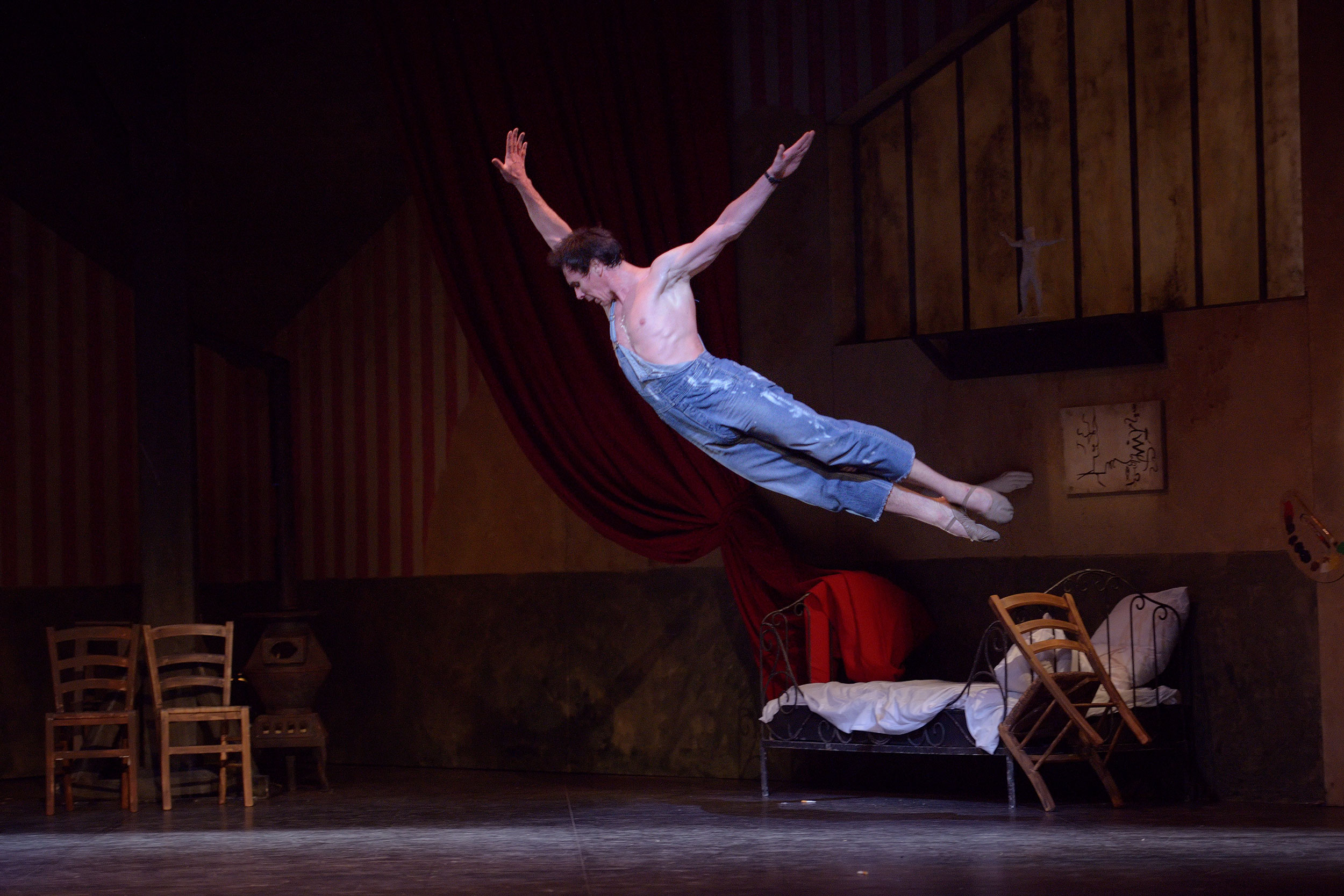 Male ballet dancer in mid-air performing an athletic leap in overalls, set against a theatrical stage background with rustic furniture and red drapery, during a performance of Le Jeune Homme et la Mort.