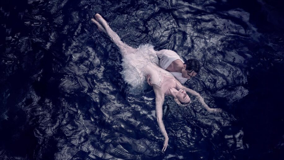 An ethereal scene from Swan Lake: a ballet dancer portraying the White Swan Odette lies gracefully in the arms of her partner, Prince Siegfried, surrounded by dark, rippling water, embodying the beauty and tragedy of the timeless ballet.