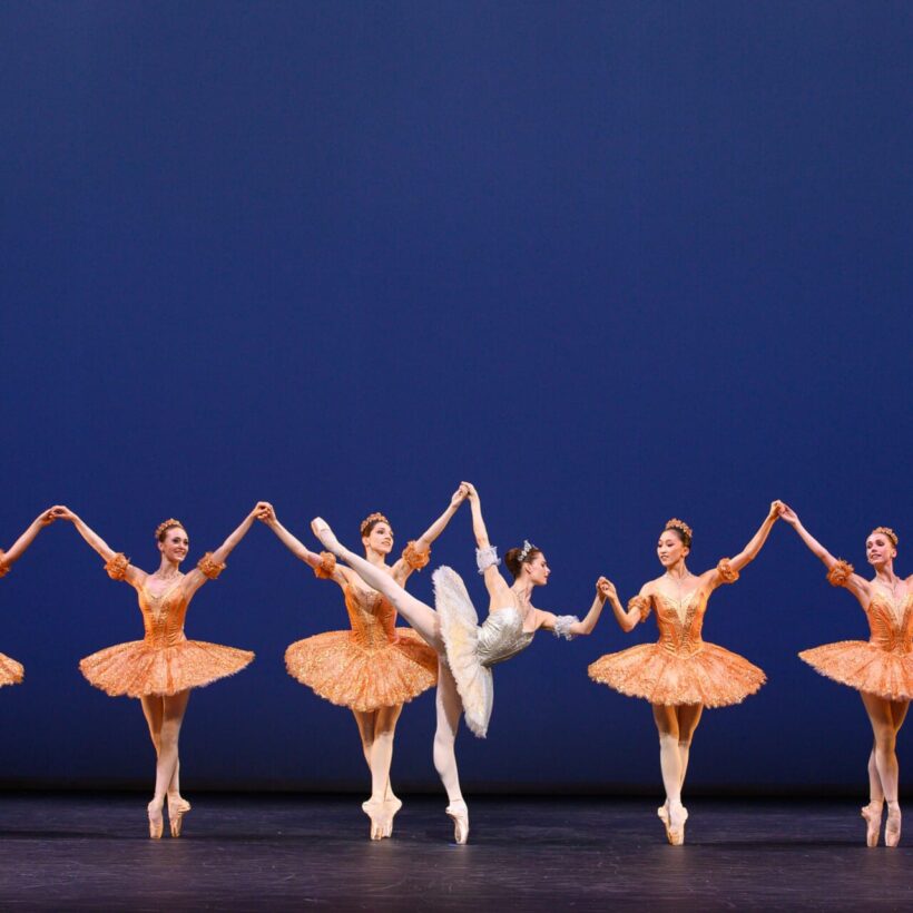 A group of ballet dancers performs on stage in matching orange tutus, with a central dancer in white striking an arabesque pose.