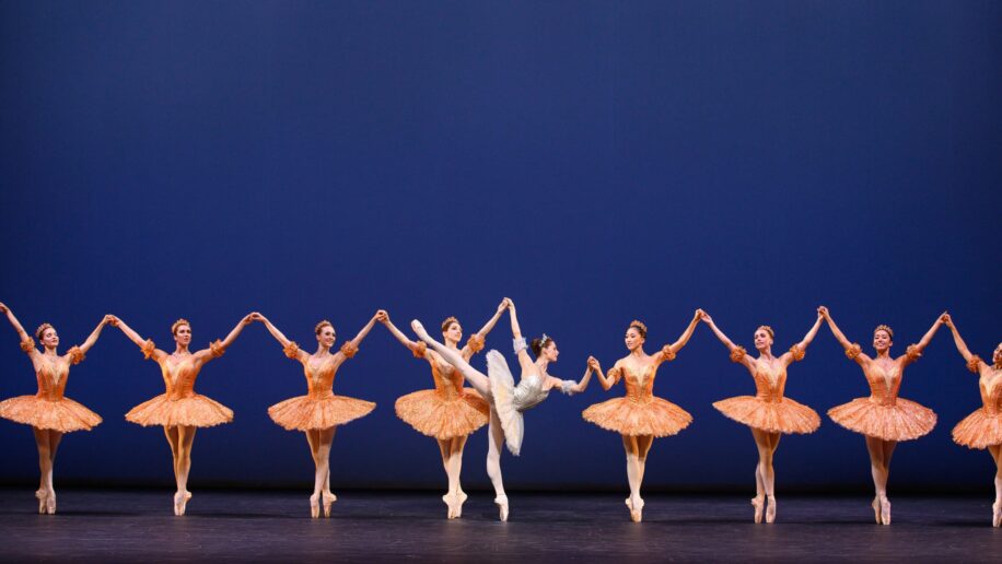 A group of ballet dancers performs on stage in matching orange tutus, with a central dancer in white striking an arabesque pose.