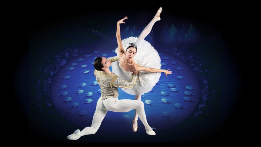 This image shows a dramatic performance of Swan Lake in-theround, featuring a male and a female dancer in an elegant pose. The male dancer, dressed in a gold-trimmed costume, is holding the female dancer, who is wearing a white tutu and tiara, as she performs a graceful arabesque. In the background there’s a circular stage with a group of dancers in white tutus laying on the ground.