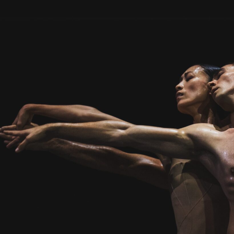 Two ballet dancers, a man and a woman, perform a deeply emotive duet against a dark background. Their bodies glisten with sweat, their arms extended in perfect unison.