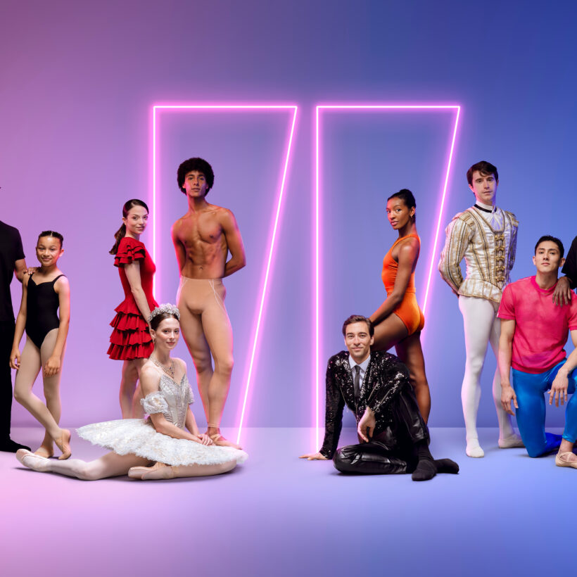 Group of dancers and creatives from English National Ballet standing and sitting against a gradient pink-to-blue background with two glowing neon rectangles. The group includes people in various ballet costumes, from classical tutus and tights to contemporary outfits and casual wear.