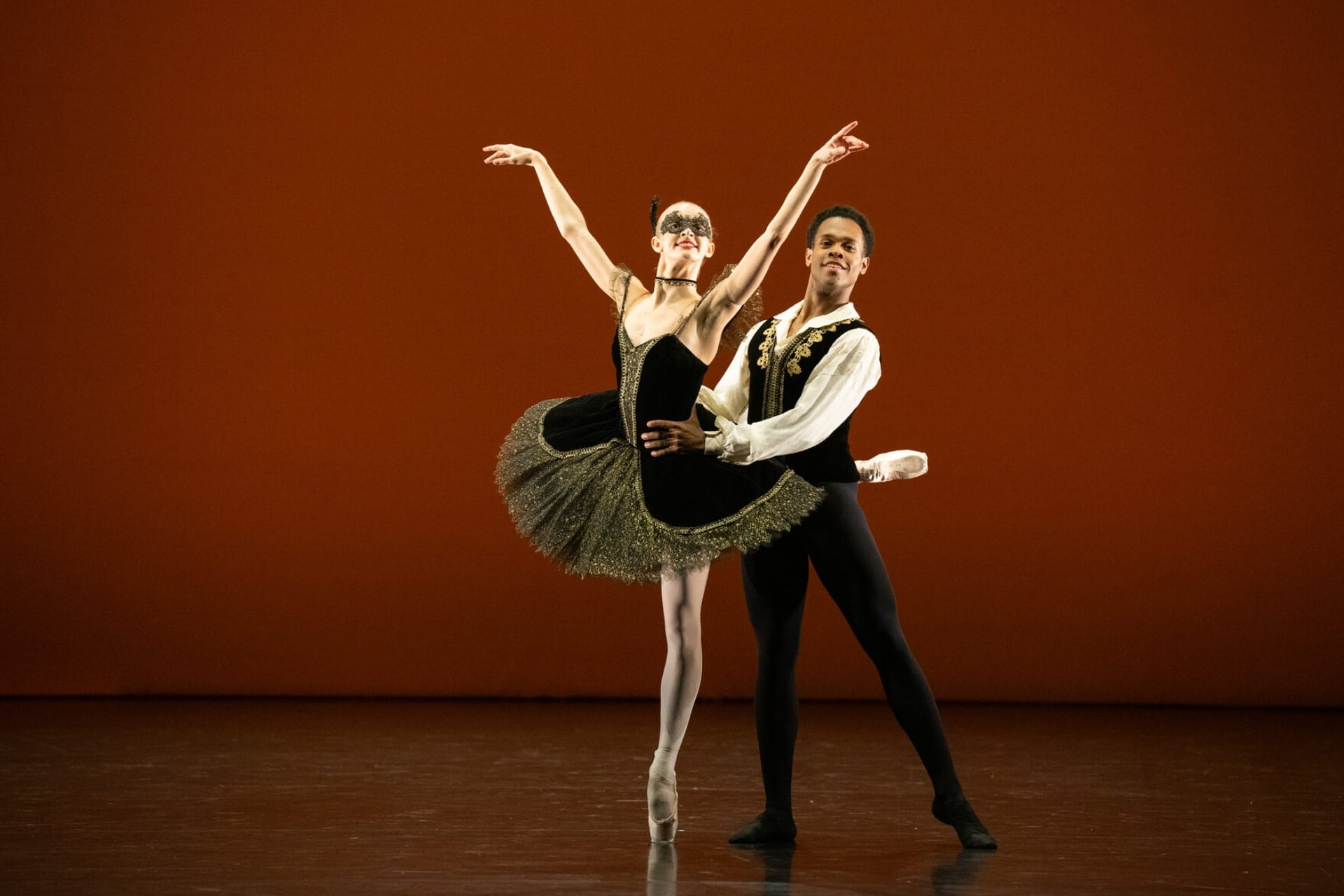 Ballet dancers performing a pas de deux on stage at Emerging Dancer 2025. A female dancer in a black tutu with gold trim executes an attitude en pointe while her male partner in a black and gold vest supports her