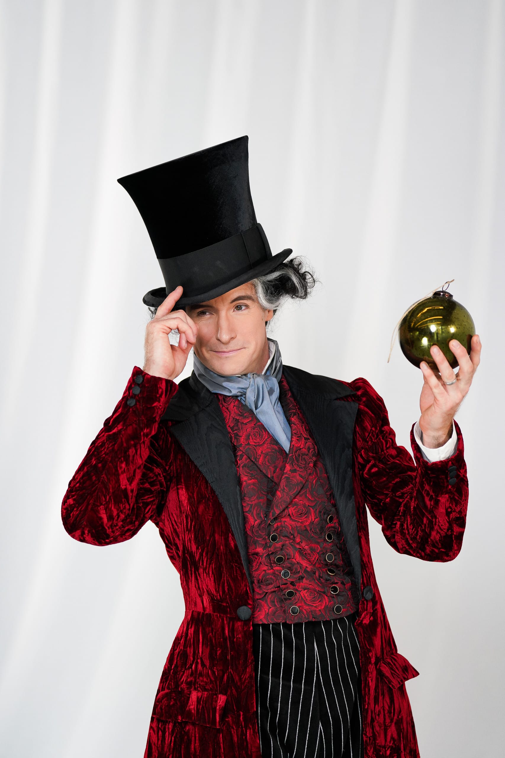 A man dressed in an elaborate costume stands against a plain white background. He wears a tall black top hat