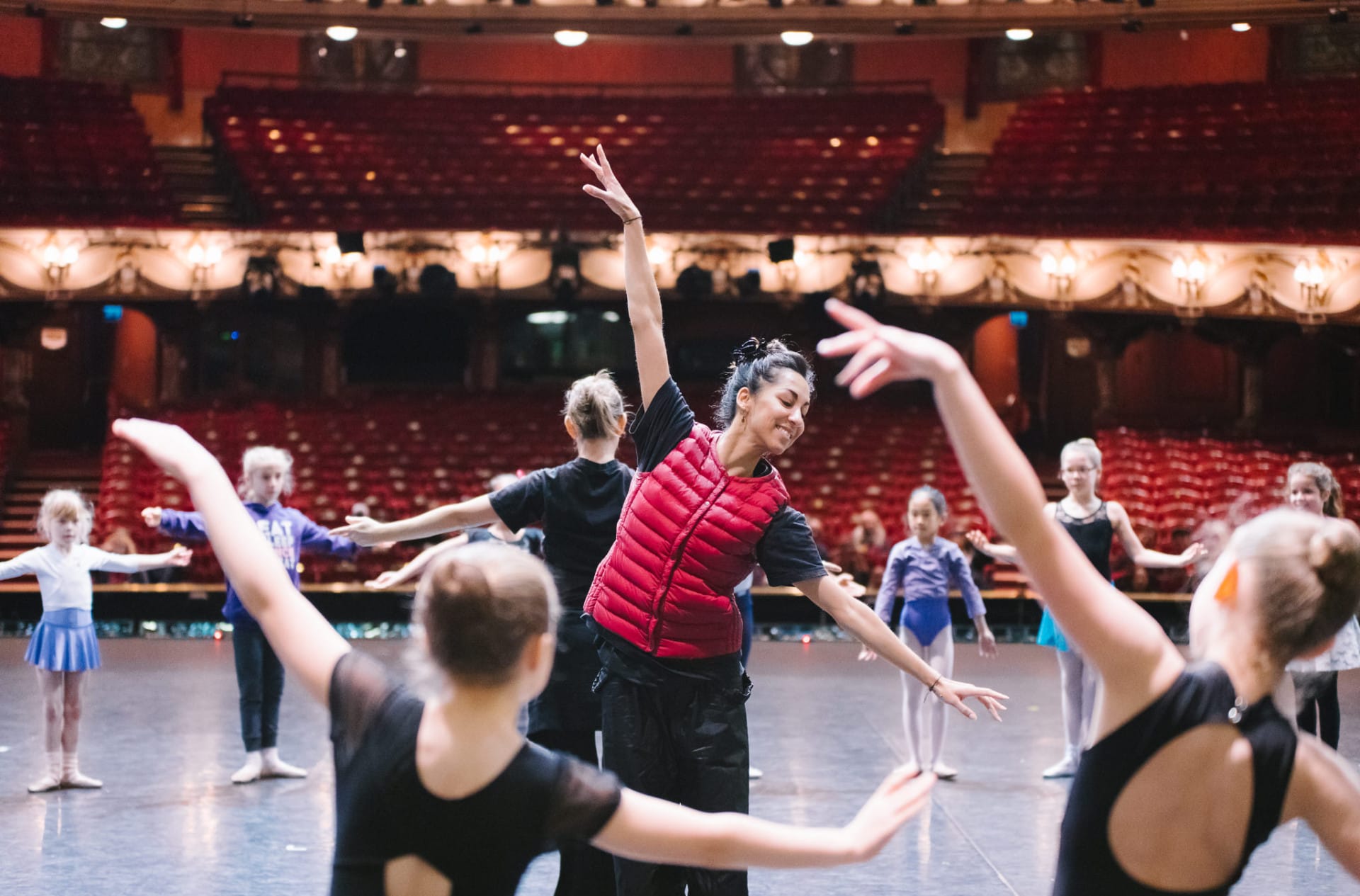 A dance artist wearing a red gilet and black trousers leads a ballet workshop for young dancers on stage at The London Coliseum. She is demonstrating a dance move that the rest of the classes is copying