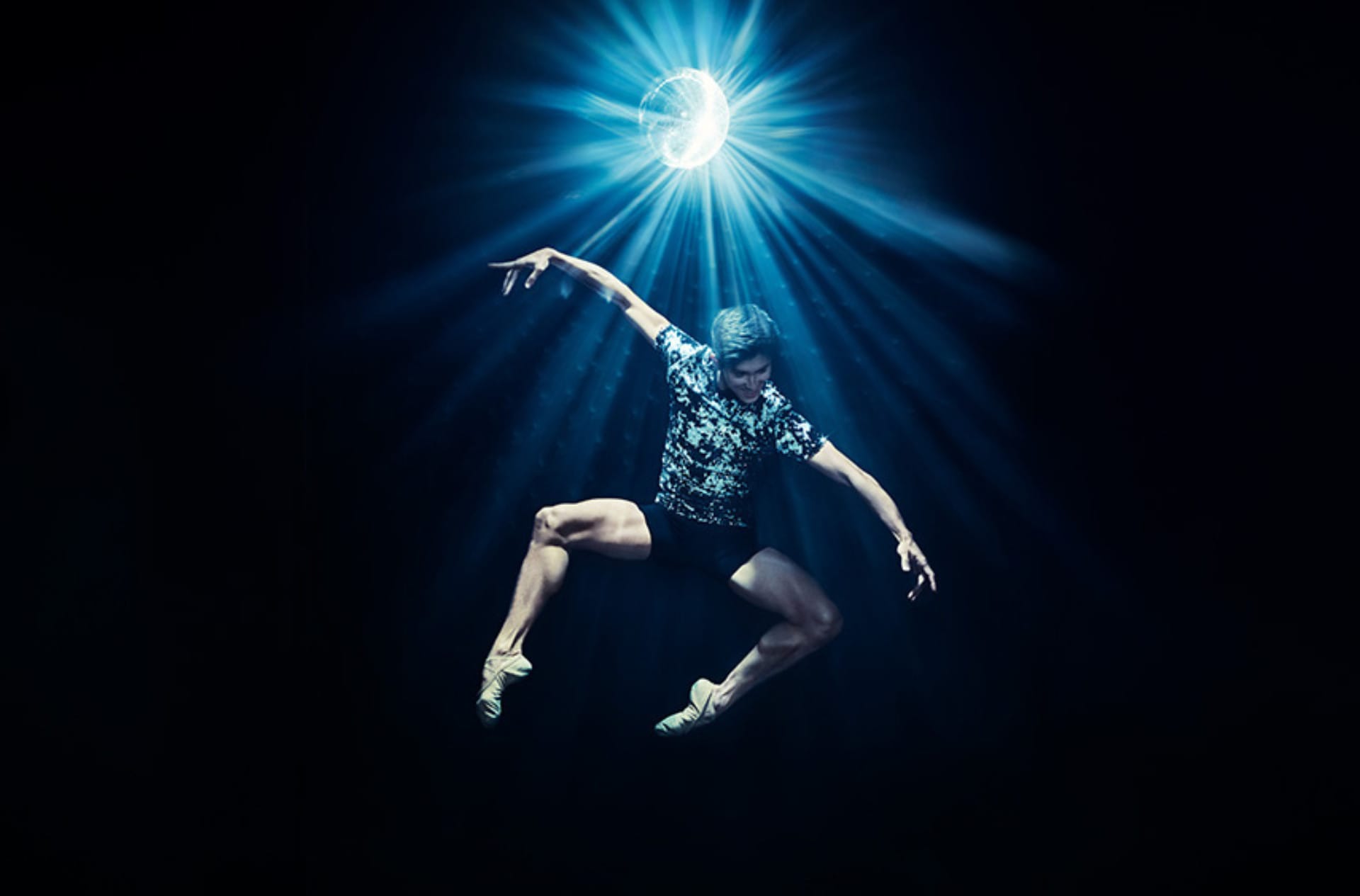 A dancer jumps in the air with their hands outstretched and knees bent underneath a disco ball reflecting light.