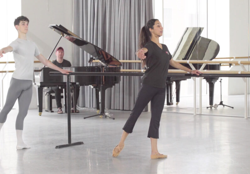 ballet teacher and a male student practice battement tendus at the barre in a bright dance studio, with a pianist playing at a grand piano in the background.