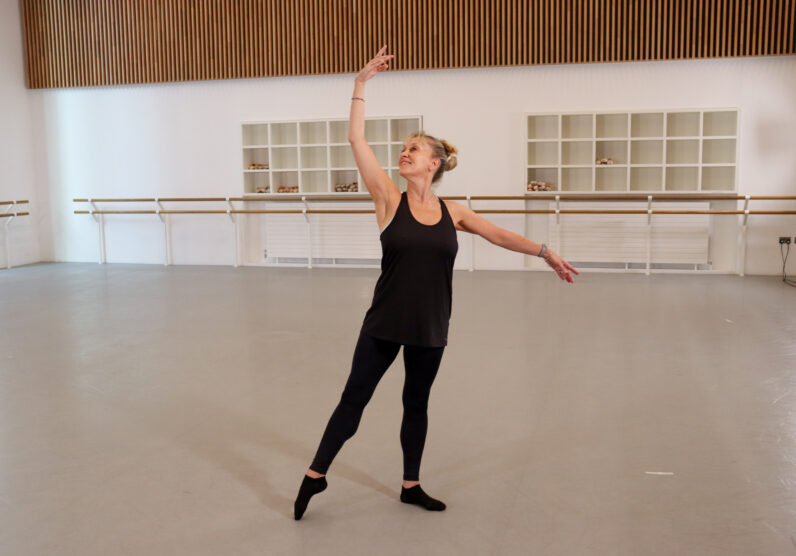 A woman in black dancewear performs a graceful ballet pose in a spacious studio, with one arm raised overhead and the other extended to the side.