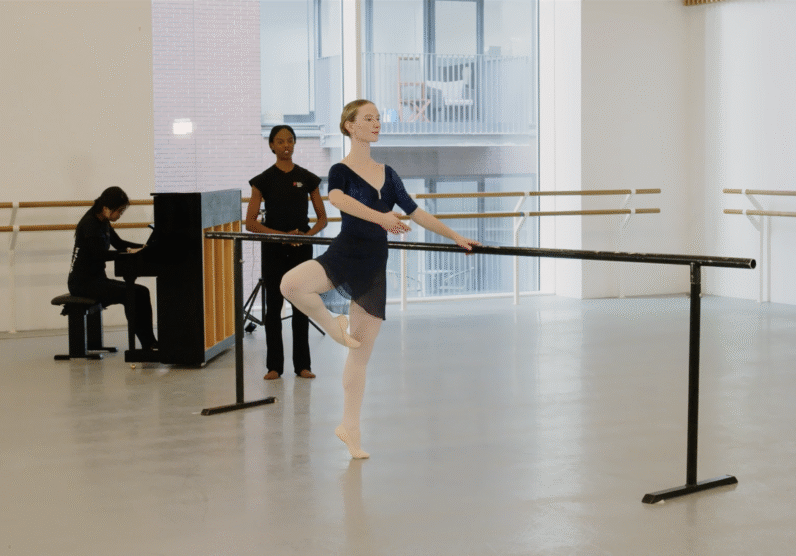 A ballet student practices at the barre in a studio, standing on one leg with the other bent in retiré position. A teacher observes nearby, while a pianist plays an upright piano in the background.