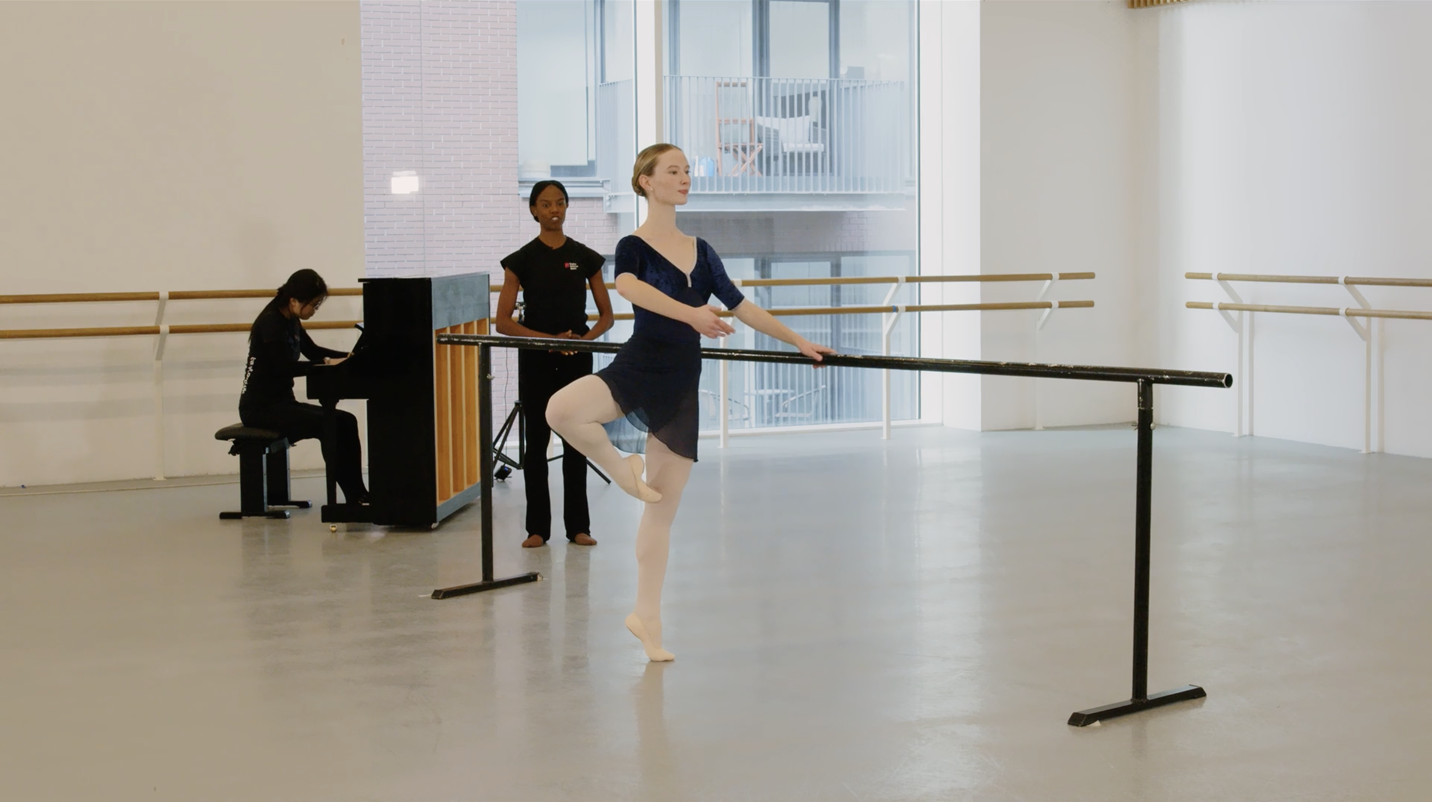 A ballet student practices at the barre in a studio, standing on one leg with the other bent in retiré position. A teacher observes nearby, while a pianist plays an upright piano in the background.