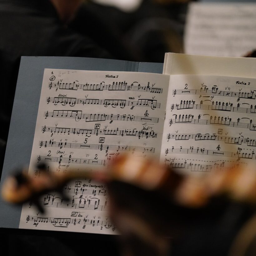 Close-up of a violinist’s music sheet during a performance or rehearsal, with handwritten notes and markings visible on the pages and a blurred violin in the foreground.