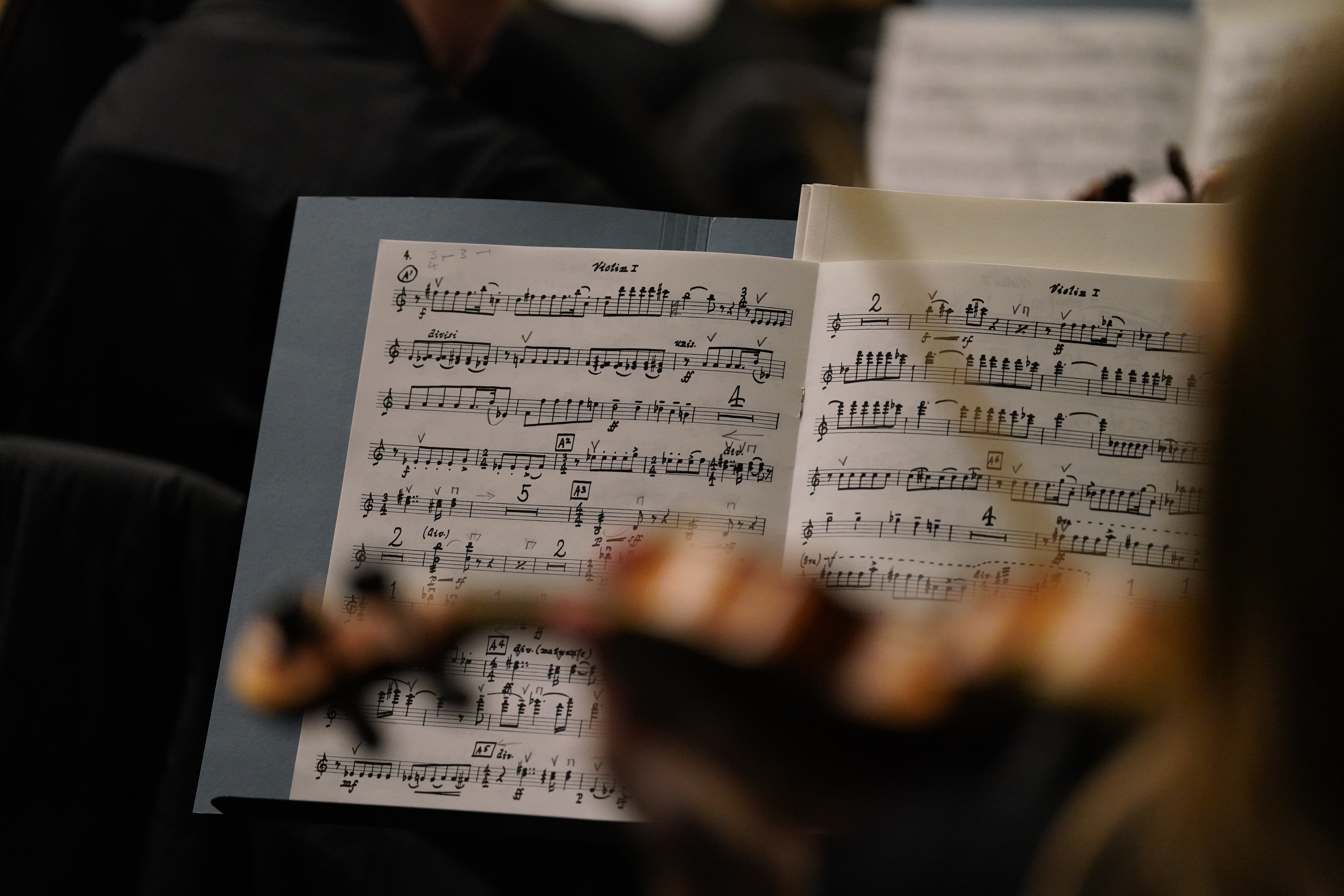 Close-up of a violinist’s music sheet during a performance or rehearsal, with handwritten notes and markings visible on the pages and a blurred violin in the foreground.