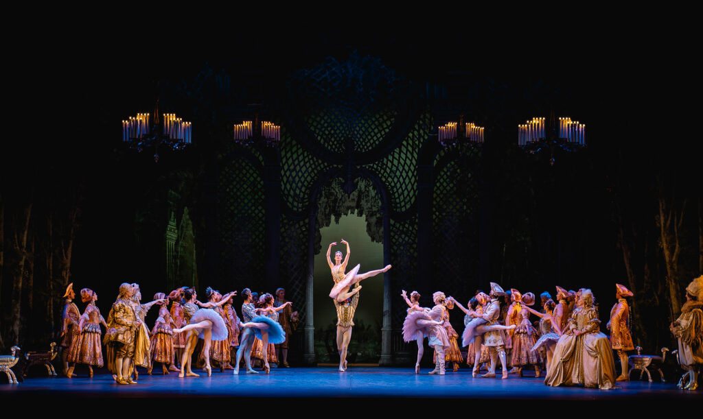 Under four chandeliers, dancers in tutus and courtly attire look towards Aurora and the Prince