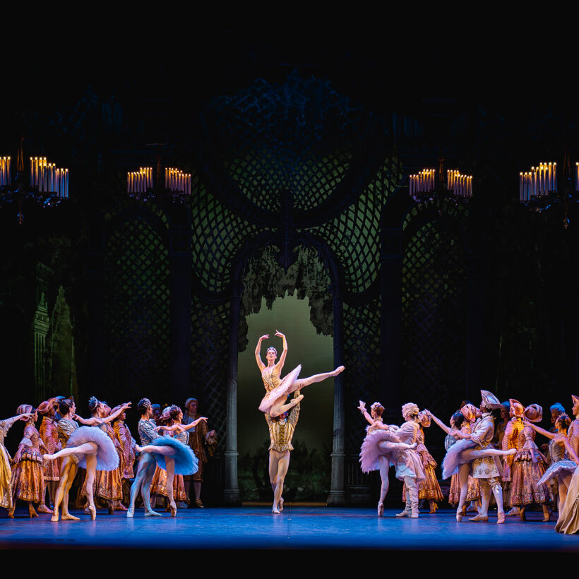 Under four chandeliers, dancers in tutus and courtly attire look towards Aurora and the Prince