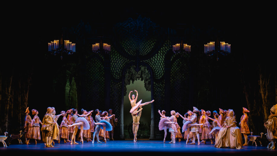 Under four chandeliers, dancers in tutus and courtly attire look towards Aurora and the Prince