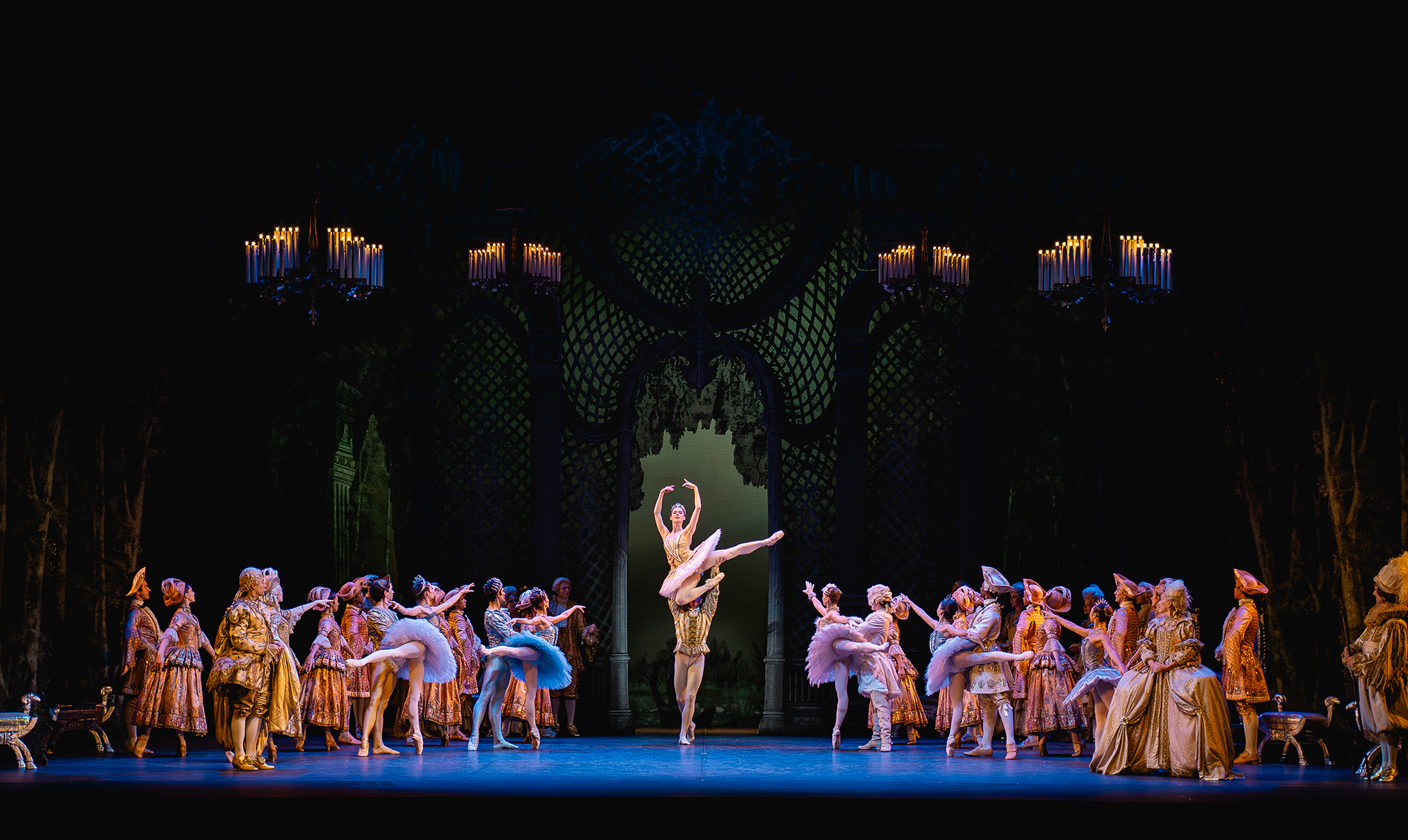 Under four chandeliers, dancers in tutus and courtly attire look towards Aurora and the Prince