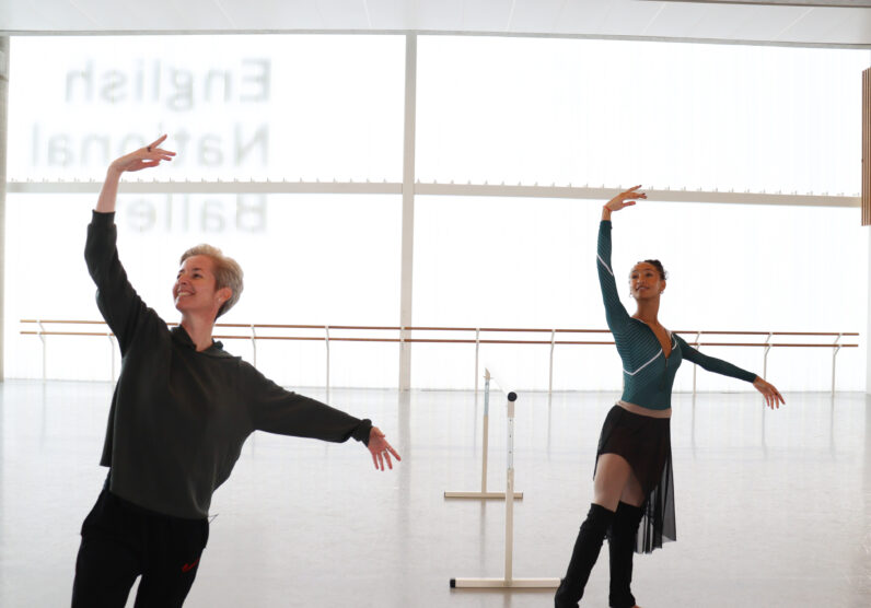 Two dancers, one older in black and one younger in teal, practice ballet with raised arms in a bright studio with large windows and a ballet barre.